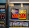 An advertisement display in a store window features lottery jackpot amounts for Mega Millions and Powerball, both showing jackpots of 208 million and 200 million respectively. Below the display, several lottery tickets and promotional posters are visible. To the left of the display, there is a refrigerator stocked with various drinks including different flavors of sports drinks and bottled water.