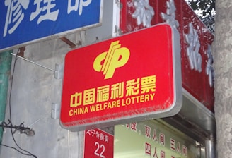 A red sign displaying the text 'China Welfare Lottery' in both Chinese and English, mounted on the exterior of a building. The surroundings include a blue sign with Chinese characters and a partial tree trunk visible on the right side.