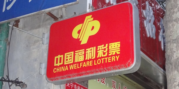 A red sign displaying the text 'China Welfare Lottery' in both Chinese and English, mounted on the exterior of a building. The surroundings include a blue sign with Chinese characters and a partial tree trunk visible on the right side.