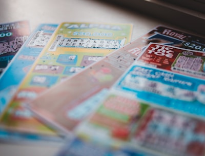 A selection of colorful lottery scratch-off tickets placed on a flat surface near a window. The image has a shallow depth of field, focusing on the tickets while blurring the background. The tickets display various games and prize amounts.