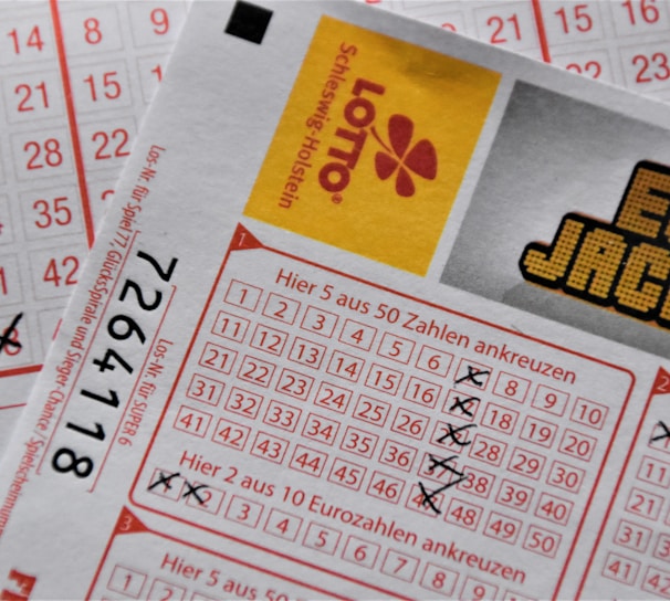 A close-up image of lottery tickets featuring crossed-out numbers, brand logos, and various numbers in a grid format. The tickets have sections for different games, and numbers are marked with black crosses.