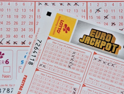 Three lottery tickets are displayed, including Eurojackpot and Super 6. Various numbers are marked on the tickets, with some crossed out and others checked. The Eurojackpot ticket is partially visible with the logo prominently displayed, and a ticket number is visible on one of the tickets.