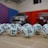 A collection of white lottery balls with black numerals scattered on a wooden surface. Two vehicles are blurred in the background, parked in a room with multicolored walls including teal, purple, blue, and red panels.