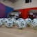 A collection of white lottery balls with black numerals scattered on a wooden surface. Two vehicles are blurred in the background, parked in a room with multicolored walls including teal, purple, blue, and red panels.