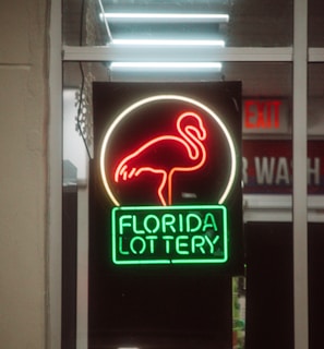 A neon sign depicting a flamingo above the words 'Florida Lottery' is prominently displayed. The sign is illuminated in bright red and green colors, and it is framed by glass panels. An 'EXIT' sign is visible in the background.