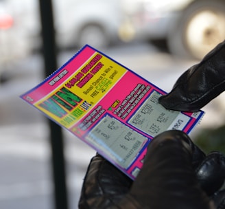 A person wearing black gloves is holding a colorful lottery scratch-off ticket. The ticket includes information about a $1,000,000 guaranteed minimum payout and features multiple games with various prize amounts.