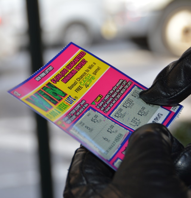 A person wearing black gloves is holding a colorful lottery scratch-off ticket. The ticket includes information about a $1,000,000 guaranteed minimum payout and features multiple games with various prize amounts.