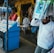 A street vendor stall displaying an array of lottery tickets clipped to a bright blue kiosk. A man in a red shirt leans on the counter, appearing thoughtful, while another man in a white shirt adjusts his hat and walks by. A variety of products hangs from strings above, and a colorful display fills the background with vibrant signs and packets. The scene suggests a busy, lively marketplace.